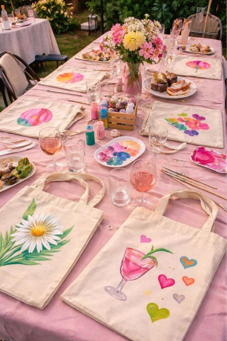 Table d’atelier dressée en extérieur avec des tote bags peints, du rosé et des fleurs